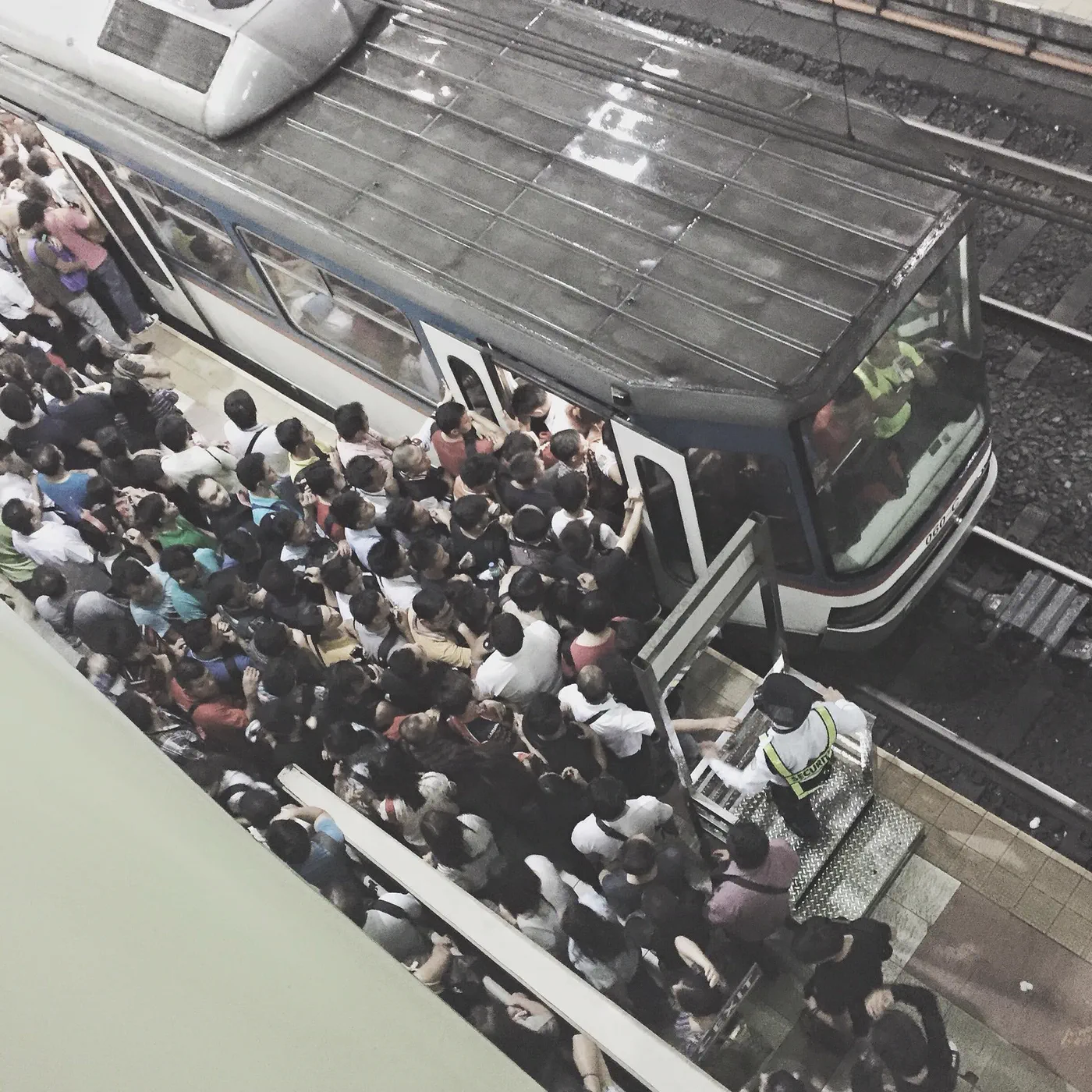 Looking down from a footbridge onto a packed station platform at rush hour, commuters pressed shoulder to shoulder around a train door while a guard watches from the steps